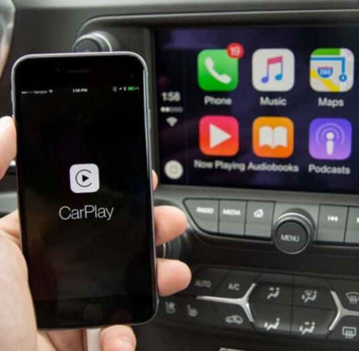 Phone running CarPlay held in front of a dash screen showing the CarPlay home grid